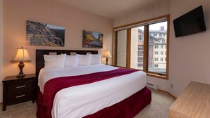 Habitación de hotel con una cama grande, dos mesitas de noche con lámparas, un televisor montado en la pared y una ventana con vistas a edificios.