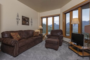 Una sala de estar con un sofá de cuero marrón, un reposapiés a juego y un sillón de cuero. Hay un televisor sobre un mueble de madera y una lámpara en una mesita auxiliar. Amplias ventanas ofrecen vistas a montañas.