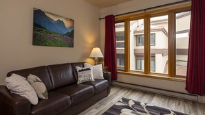 Un salon avec un canapé en cuir marron, un tableau représentant des montagnes, une lampe et une vue sur des immeubles à travers de grandes fenêtres ornées de rideaux rouges.
