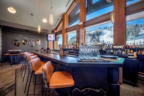 Uma área de bar com um balcão comprido, banquetas e vista para uma paisagem nevada através de grandes janelas.