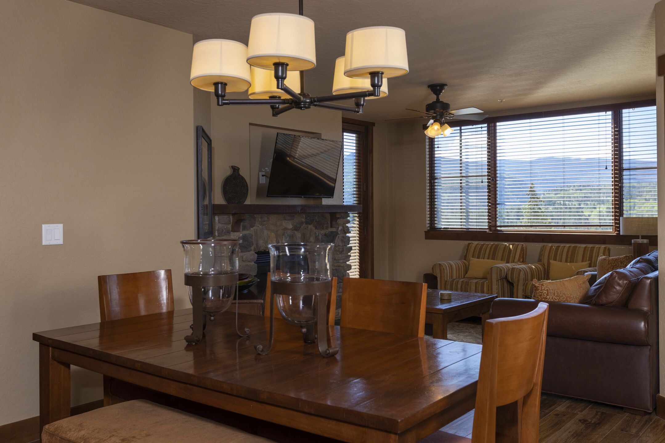 A dining area with a wooden table, chairs, and a chandelier. There is a television on the wall and a window with blinds.