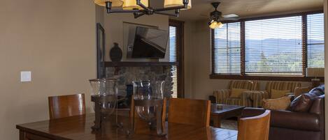 A dining area with a wooden table, chairs, and a chandelier. There is a television on the wall and a window with blinds.