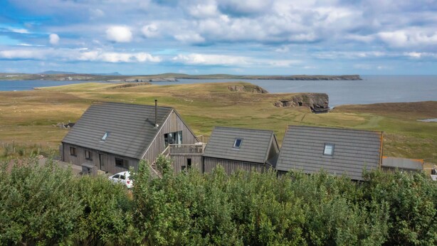 Een kustpand met drie houten gebouwen, een auto en uitzicht op de zee en heuvels.