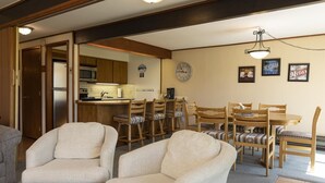 Un salon chaleureux avec une cheminée, un coin repas avec des chaises et une table en bois, et une cuisine en arrière-plan.