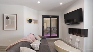 Una sala de estar moderna con un sofá gris, una mesa de centro de madera, un televisor de pantalla plana sobre la chimenea y un ventanal corredero que da a un patio exterior.