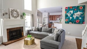 Ein Wohnzimmer mit Kamin, grauen Sofas, einem runden Couchtisch und einer Küche im Hintergrund.