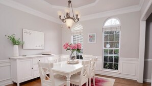 Ein Esszimmer mit einem weißen Tisch, Stühlen und einer Chandelüstere. Ein Fenster mit Jalousien und eine Vase mit Blumen stehen auf dem Tisch.