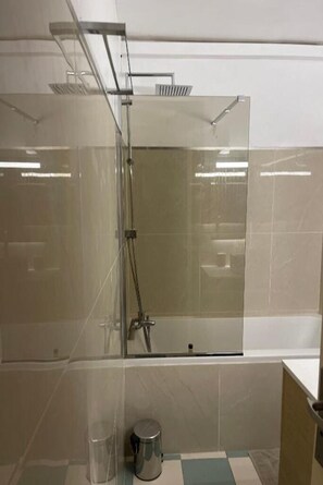 A modern bathroom with a glass-enclosed shower, a small trash bin, and a tiled floor.