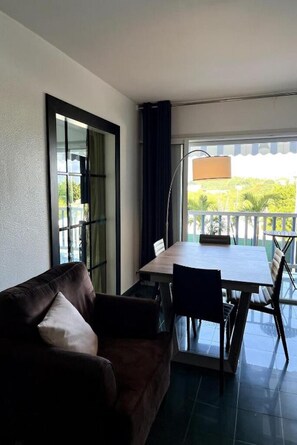 A room with a brown leather sofa, a wooden dining table with chairs, a floor lamp, and a view of greenery outside.