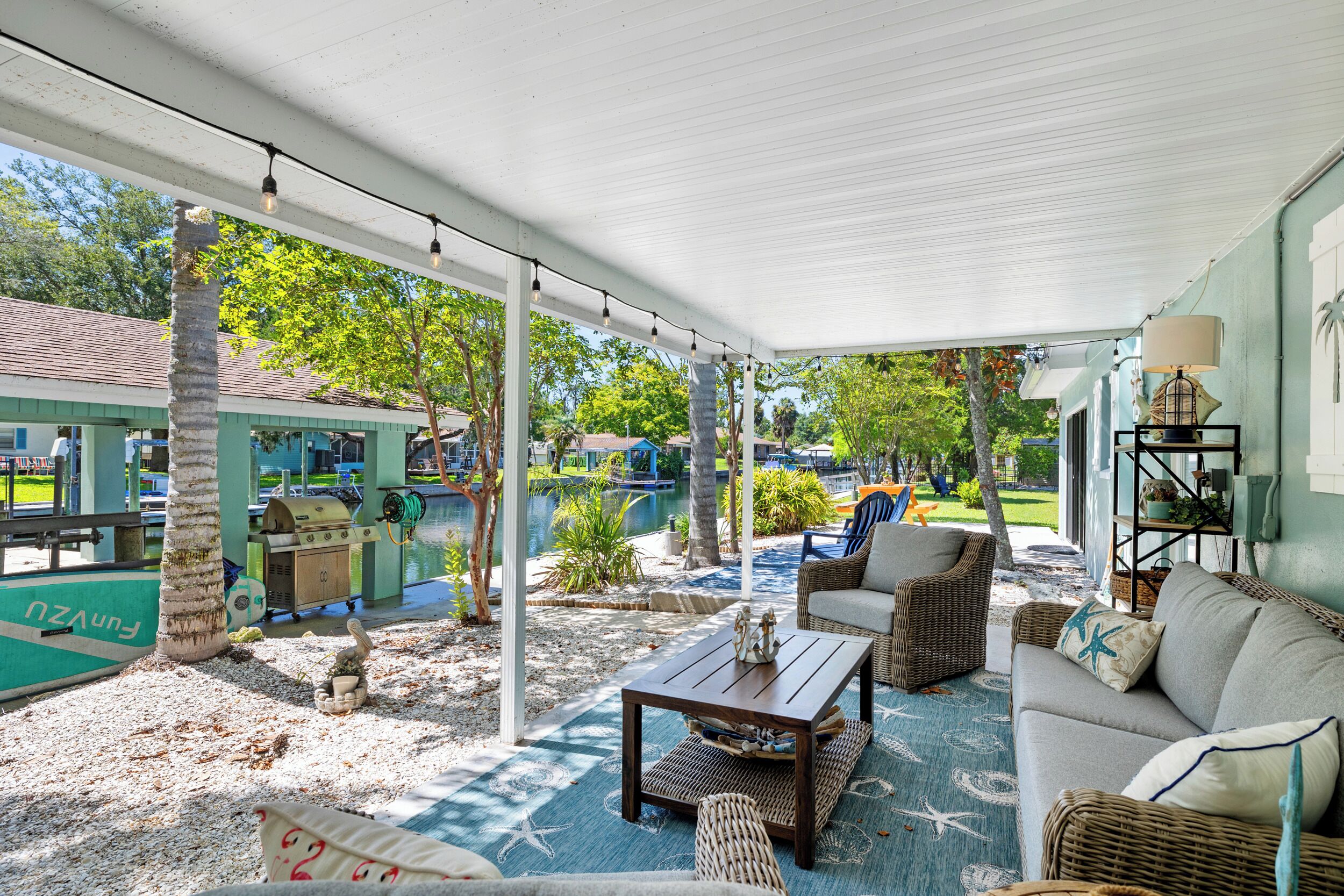 Un espace extérieur aménagé en terrasse, doté d’un coin salon couvert, d’un barbecue et offrant une vue sur un plan d’eau.