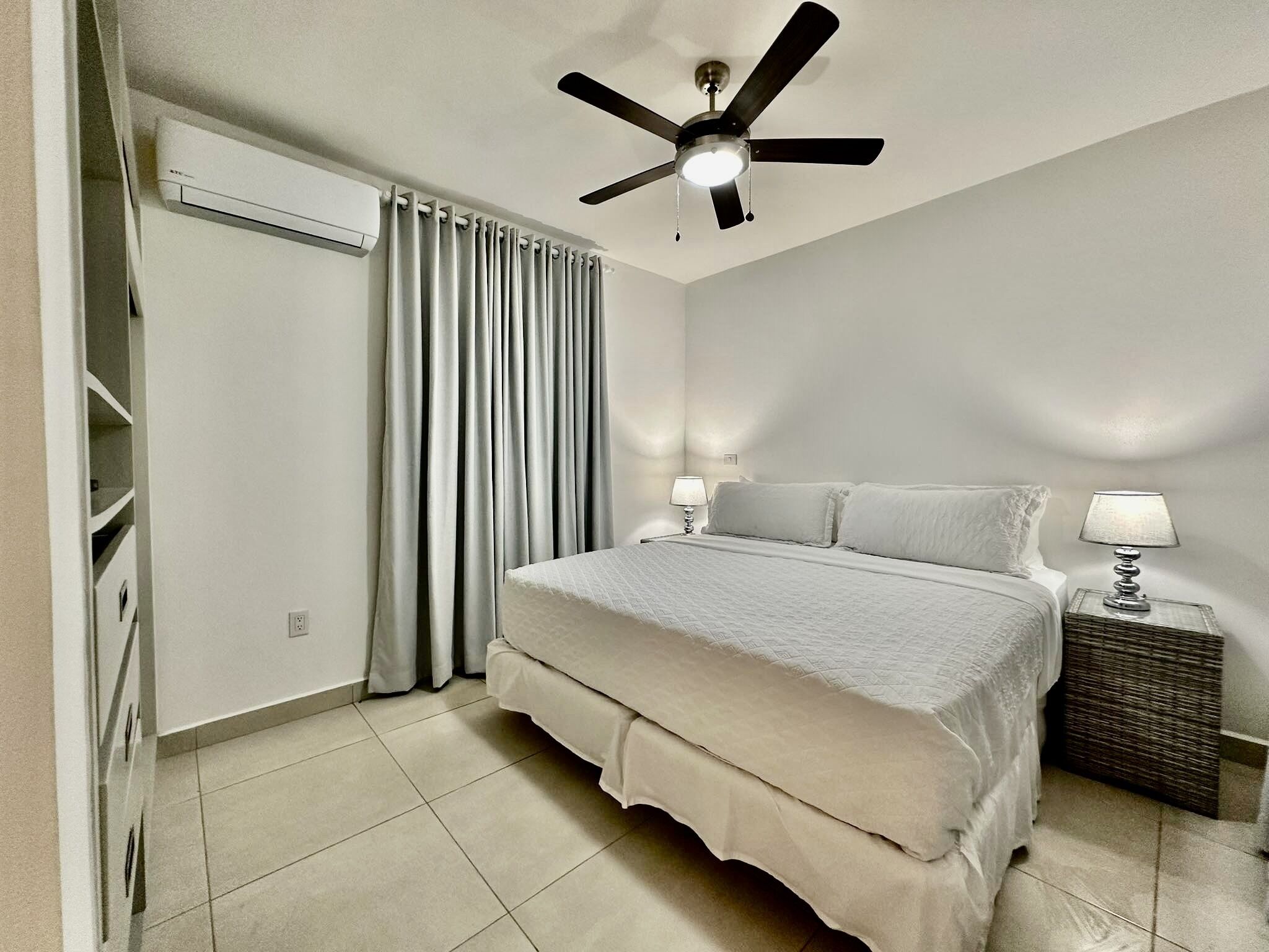 A bedroom with a bed, a ceiling fan, a nightstand with a lamp, and a window with curtains.