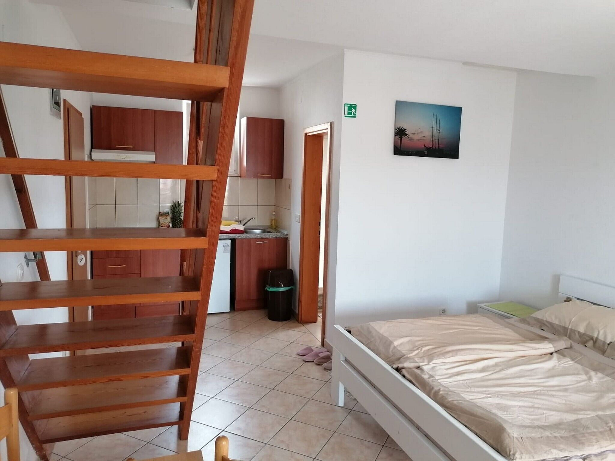 Une chambre avec un escalier en bois, un lit, une petite kitchenette et un tableau accroché au mur.