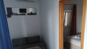 Habitació petita amb un sofà, una taula baixa, un llit i un televisor fixat a la paret.