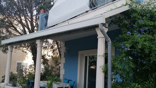 Una casa de dues plantes amb un porxo cobert, plantes en testos i una porta blava.