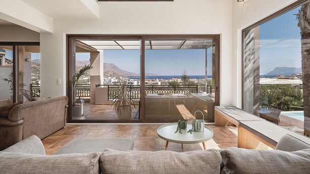 A spacious living room with a large sofa, a small round table, and a balcony with a view of the city and mountains. - Villa Anemeli | Infinity pool ,sea view close to the city, Elite Crete II (Kissamos)