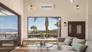 A spacious living room with a large window offering a view of palm trees and a distant landscape. - Villa Anemeli | Infinity pool ,sea view close to the city, Elite Crete II (Kissamos)
