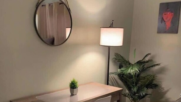 Ein Zimmer mit einem runden Spiegel, einem Schreibtisch mit einer kleinen Pflanze, einer Stehlampe und einem Blumentopf.