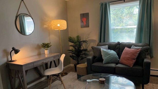 Un salon comprenant un bureau, une table basse ronde en verre, un canapé avec des coussins, un fauteuil, une lampe et une plante en pot.