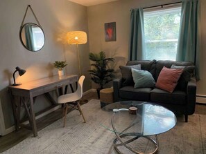 A living room with a desk, a round glass coffee table, a sofa with cushions, a chair, a lamp, and a potted plant. - Apt 4 Convenient Location (Masury)