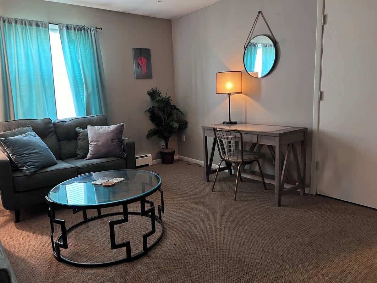 Un salon comprenant un canapé, une table basse en verre, un bureau avec une chaise, une lampe et une plante en pot.