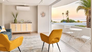 En moderne stue med en sofa, en gul stol og et spisebord på en balkon med udsigt over en strand.