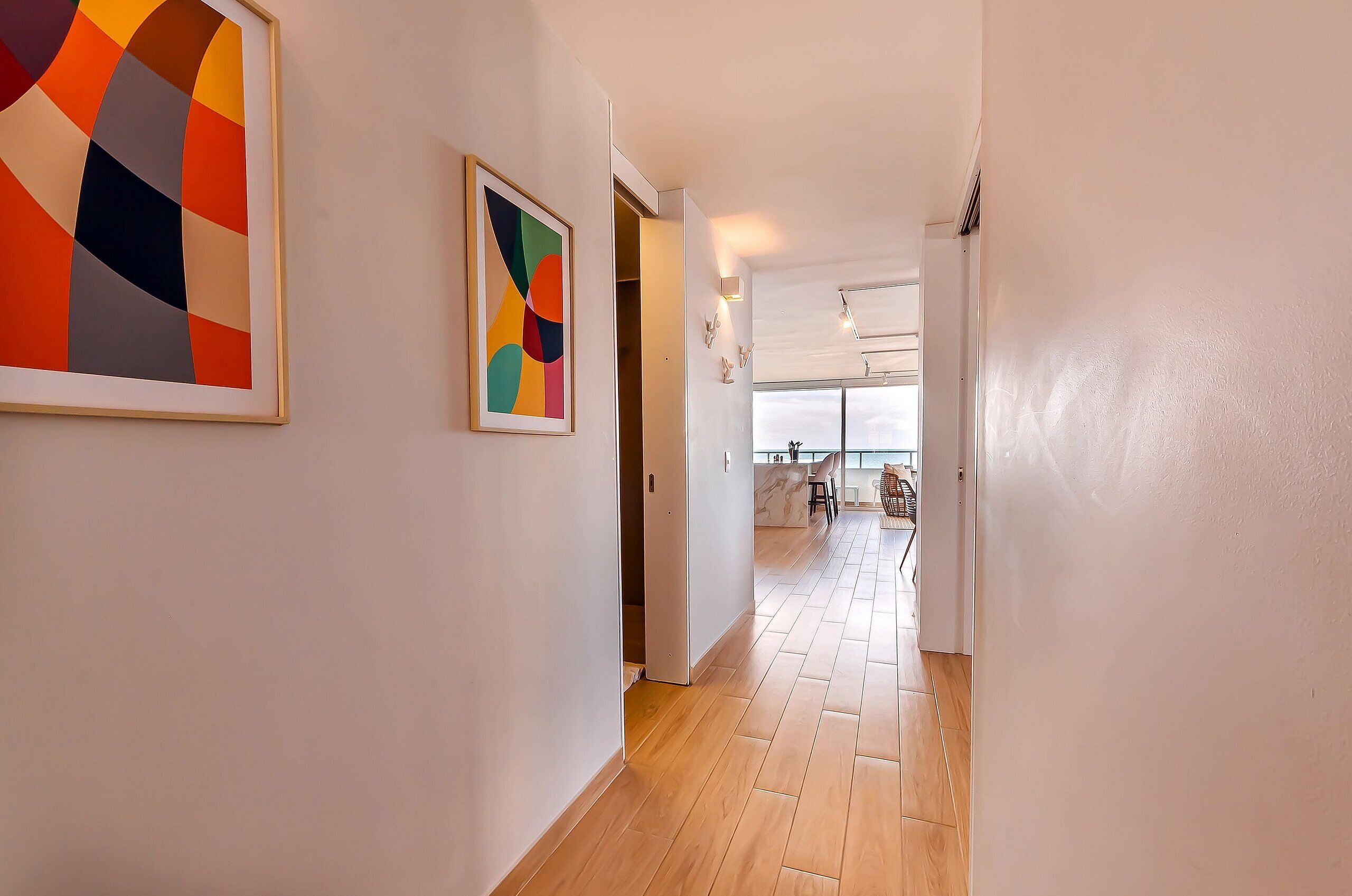 A hallway with wooden flooring, a door leading to a room with a view, and two framed artworks on the walls.