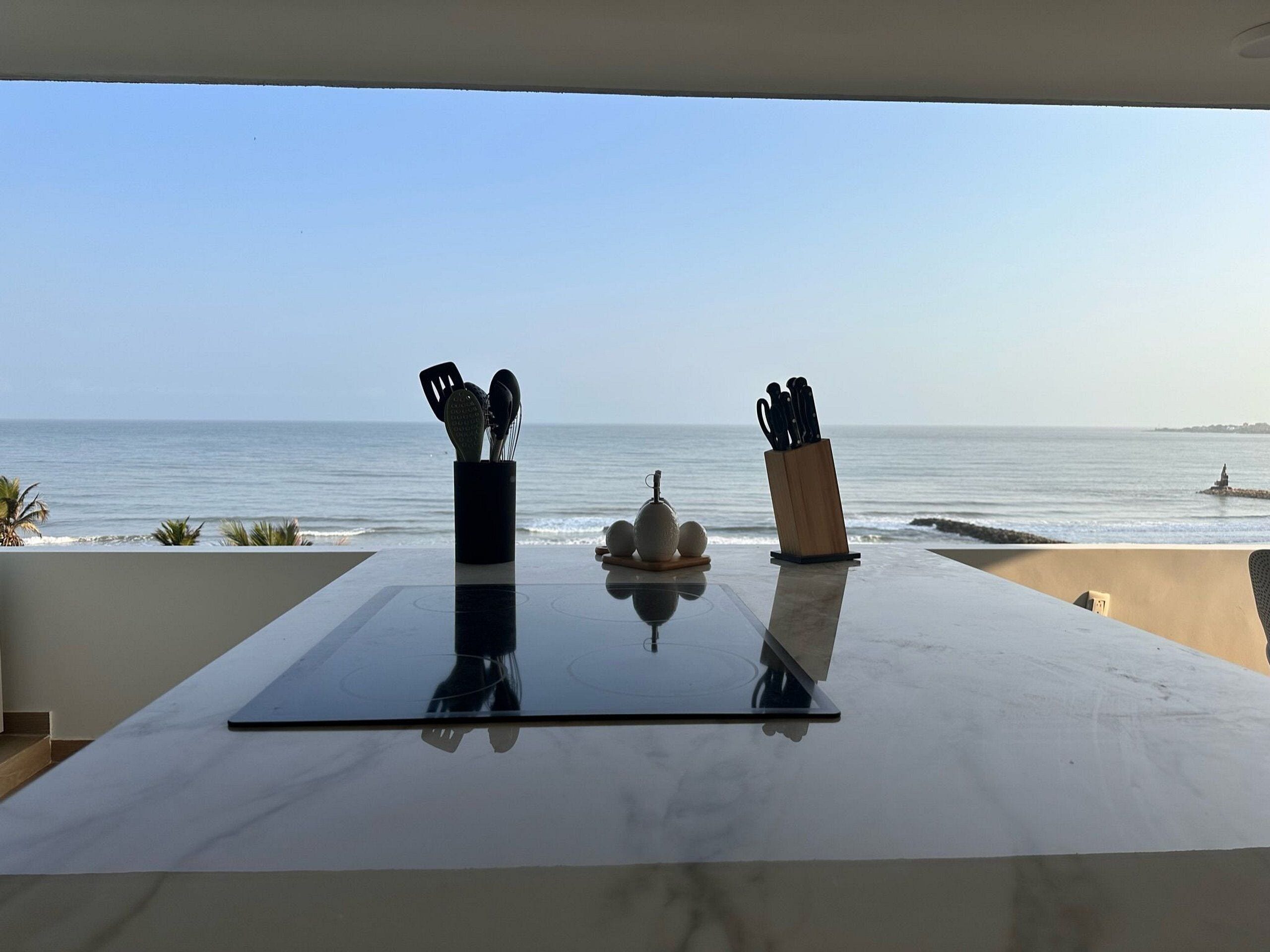 A table with kitchen utensils and a decorative item, overlooking a beach with calm waters and a clear sky.