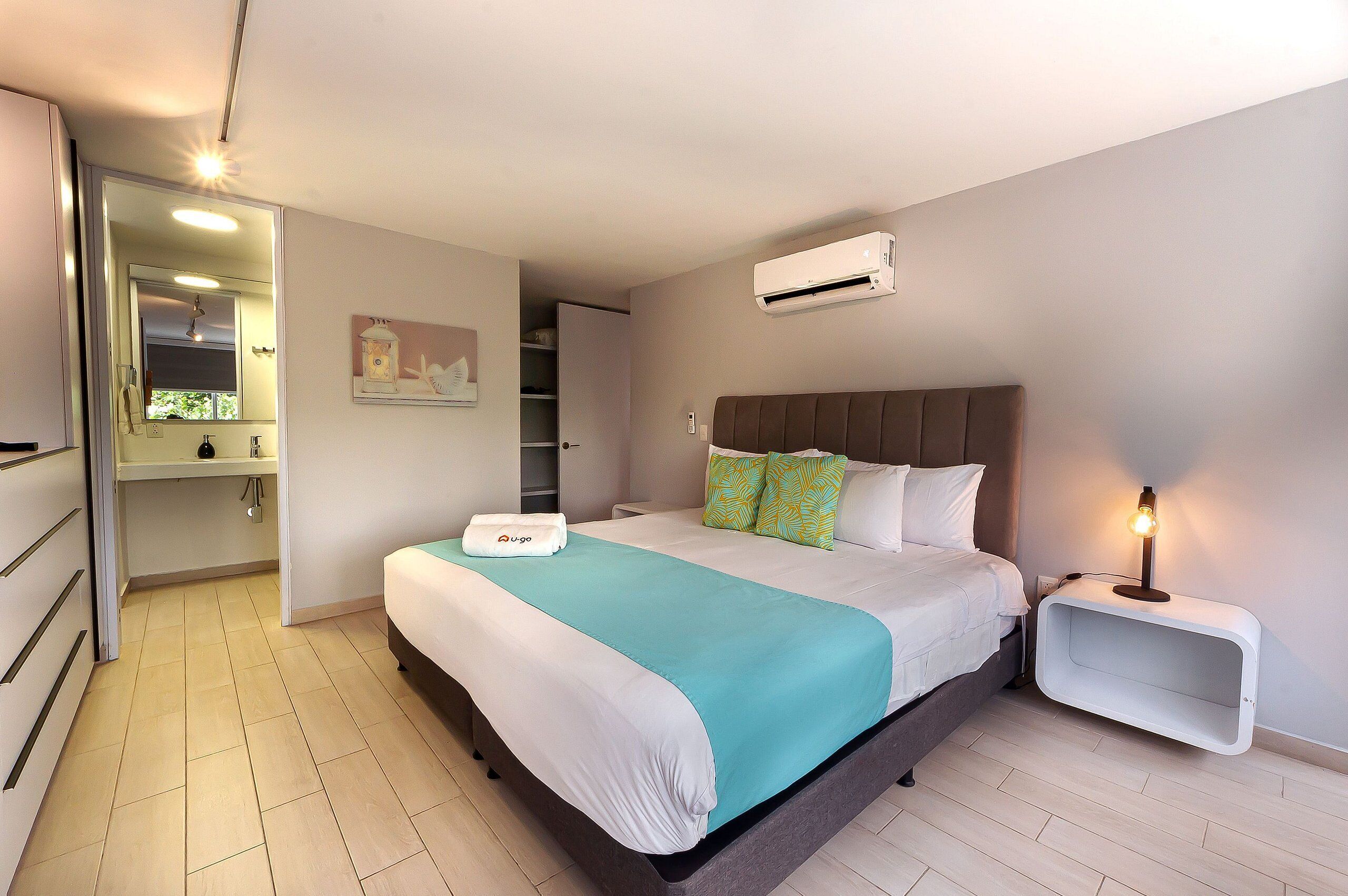 A hotel room with a bed, a bedside table with a lamp, an air conditioning unit, and a bathroom with a shower and toilet.