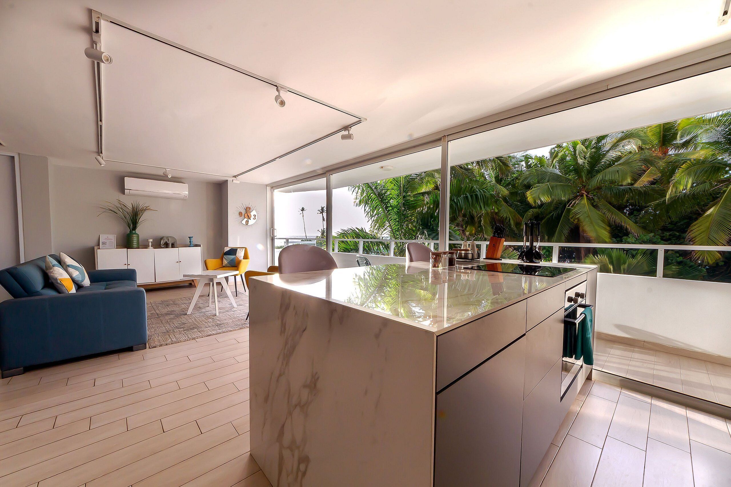 A modern kitchen with a large island, a dining area with a blue sofa, and a view of tropical foliage through large windows.