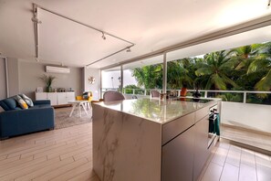 Une cuisine moderne dotée d’un grand îlot central, d’un coin repas avec un canapé bleu et d’une vue sur une végétation tropicale grâce à de grandes fenêtres.
