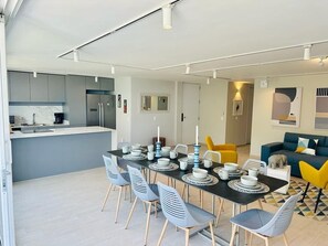 Une cuisine moderne dotée d’un réfrigérateur encastré, un coin repas avec une table noire et des chaises grises, et un salon avec un canapé bleu et des fauteuils jaunes.