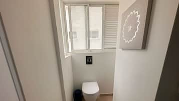 Un baño estrecho con un inodoro, una ventana con persianas y un interruptor de luz en la pared.