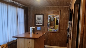 Un intérieur en bois comprenant un coin bar, une chaise, une petite table, un tableau encadré et une fenêtre avec des rideaux.