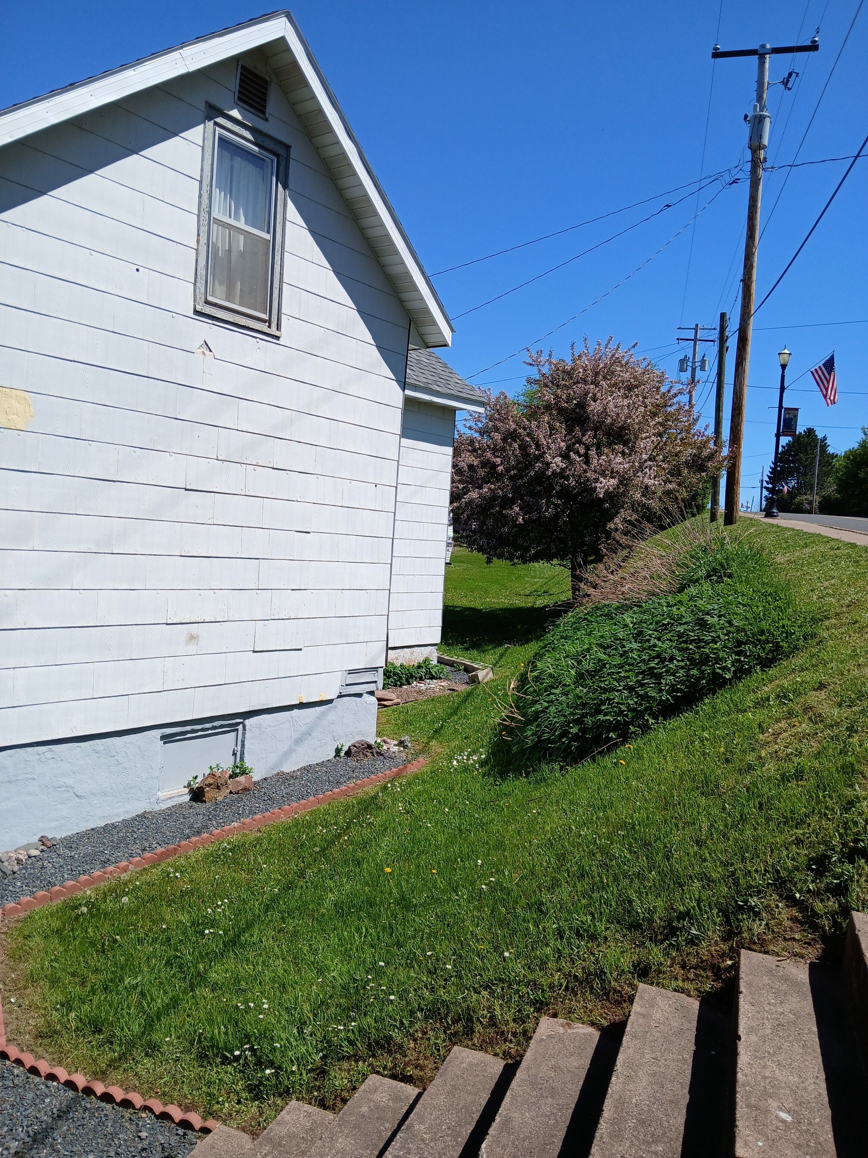 A residential house with a sloped roof, a window with a curtain, a small garden with a tree, and a sidewalk leading to a street.