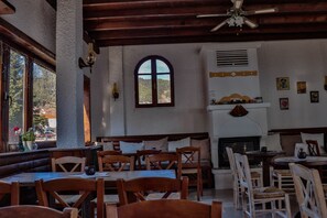 A cozy restaurant with wooden tables and chairs, a fireplace, and a large arched window overlooking a scenic view. - Hotel Mitikas (Elassona)