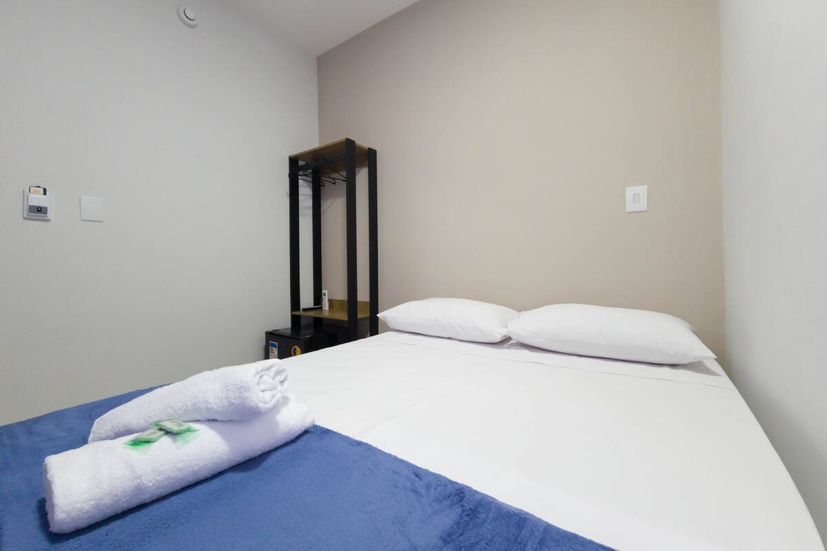 Quarto de hotel com cama, manta azul, almofadas brancas, toalha branca, prateleira preta e interruptor de luz fixado na parede.