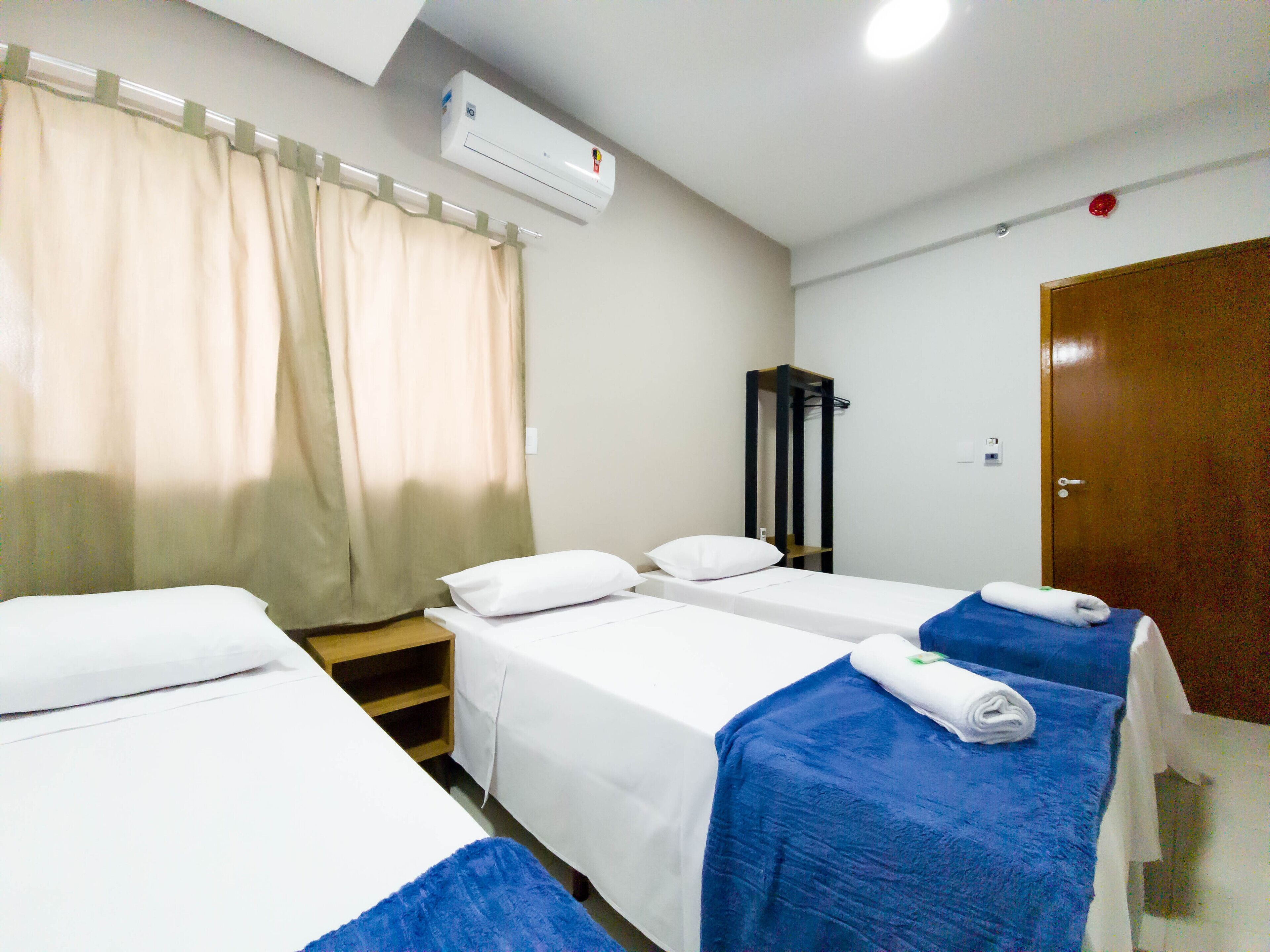 Quarto de hotel com duas camas de solteiro, um guarda-roupa de madeira, uma janela com cortinas bege e um aparelho de ar condicionado na parede.