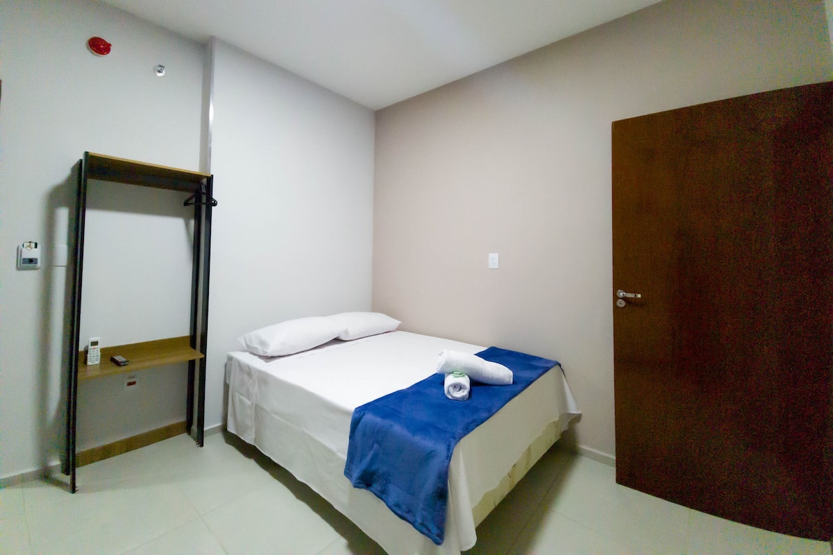 Quarto pequeno e limpo com uma cama de solteiro, uma prateleira de madeira, um espelho e uma porta.