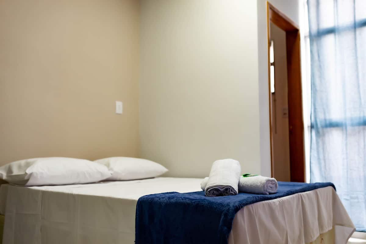 Quarto de hotel com cama, travesseiros brancos, manta azul e toalha enrolada na cama.