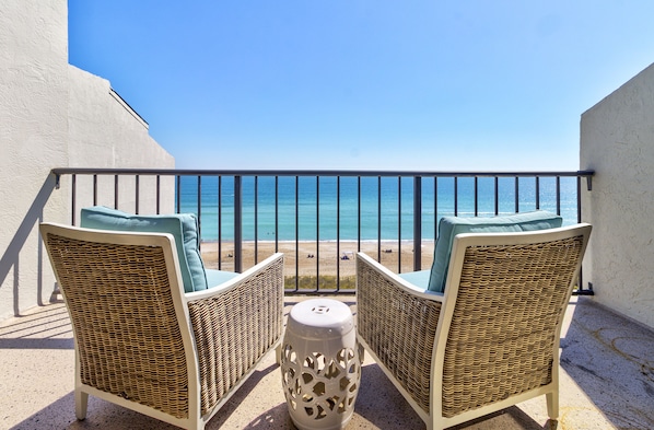 Ein Balkon mit Korbstühlen und einem kleinen Tisch, mit Blick auf einen Strand und den Ozean.