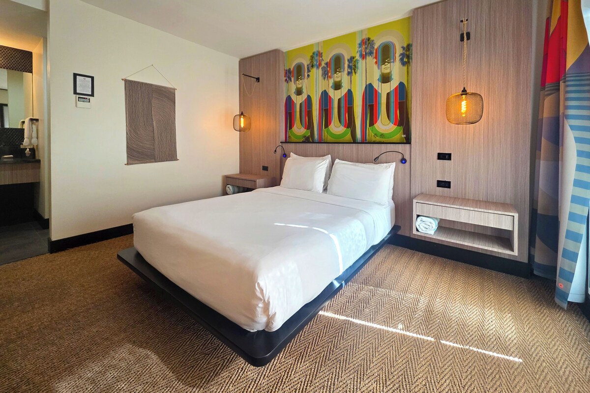 Une chambre d’hôtel moderne avec un grand lit, une tête de lit ornée d’une fresque colorée, une table de chevet avec une lampe et une petite étagère.