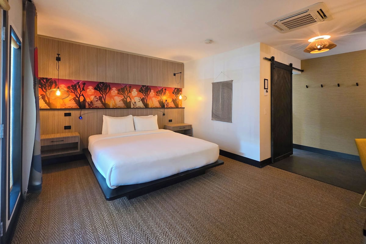 A hotel room with a bed, a headboard with a painting, a wall-mounted air conditioner, and a sliding door.