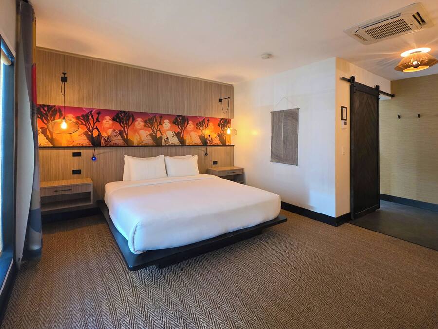A hotel room with a bed, a headboard with a painting, a wall-mounted air conditioner, and a sliding door.