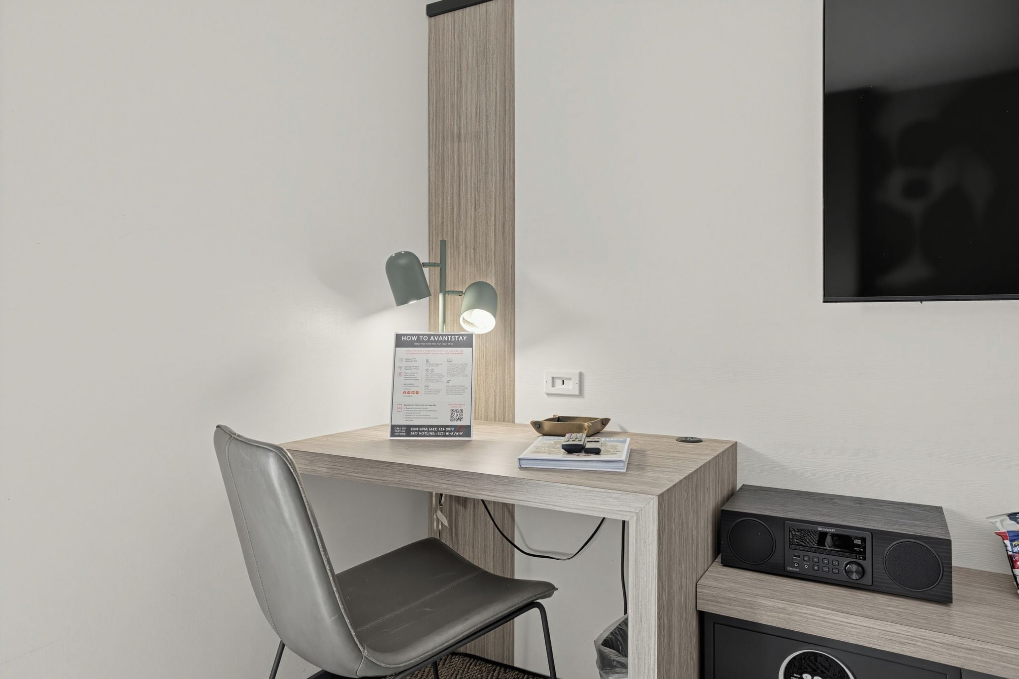 Quarto de hotel com escrivaninha, cadeira, luminária, placa e rádio.