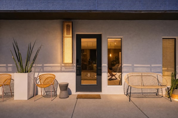 A modern exterior with a large planter, two wicker chairs, a small table, and a door with a number '6' visible inside.