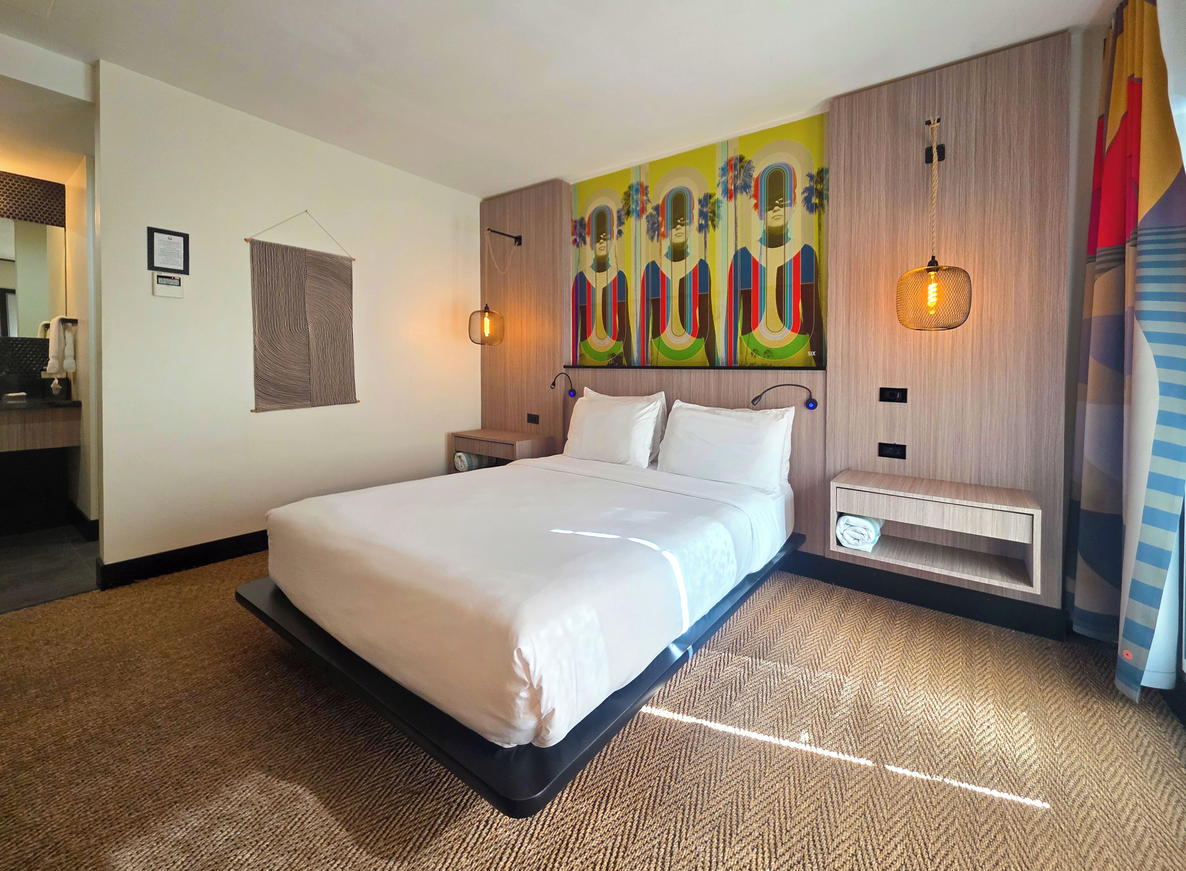 Una habitación de hotel moderna con una cama grande, un cabecero con un mural colorido, una mesita de noche con una lámpara y un estante pequeño.