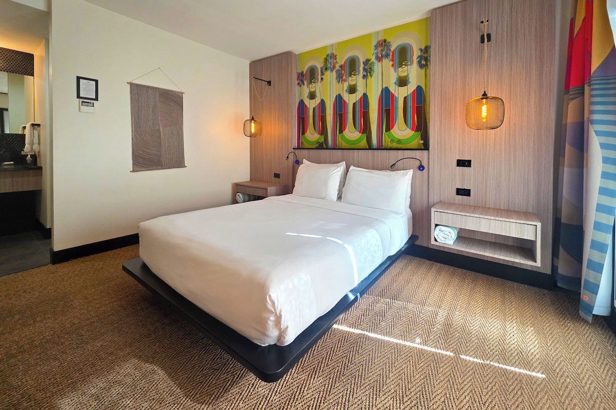 Ein modernes Hotelzimmer mit einem großen Bett, einem Kopfteil mit Wandgemälde, einem Nachttisch mit Lampe und einem kleinen Regal.