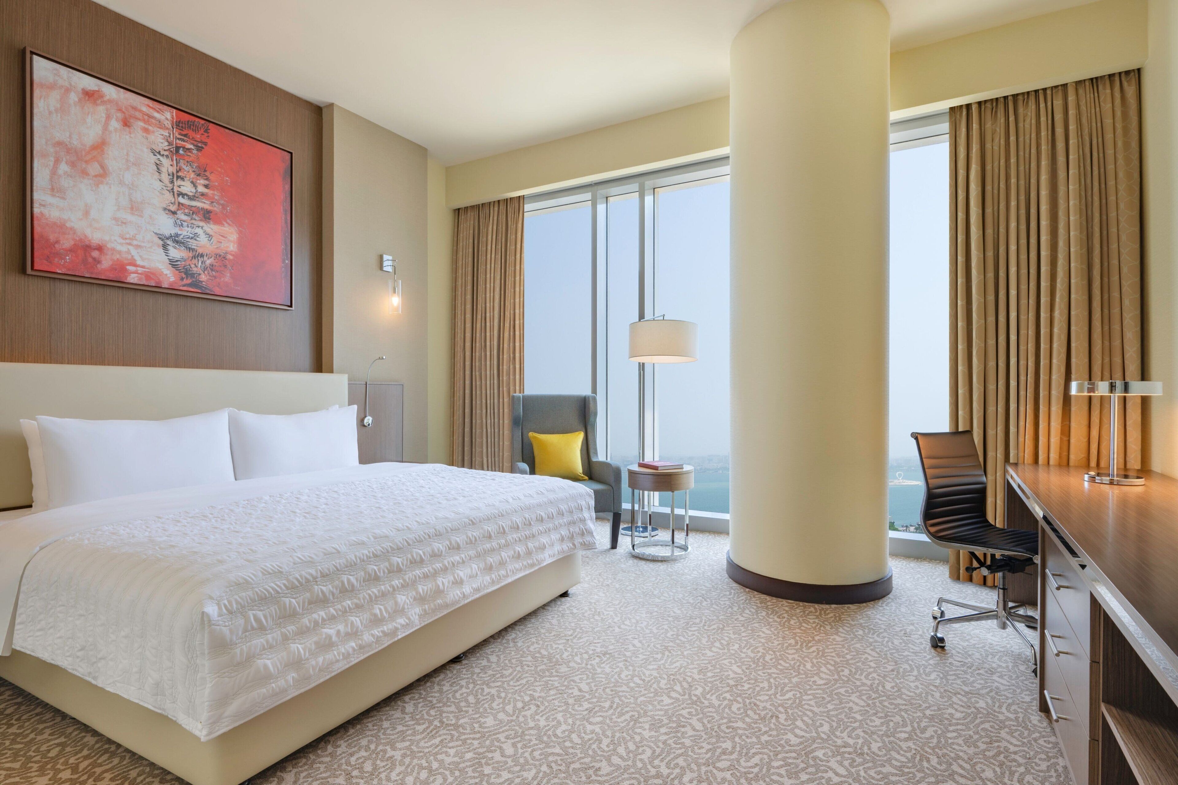 A hotel room with a large bed, a desk, a chair, a lamp, and a view of the city through large windows.