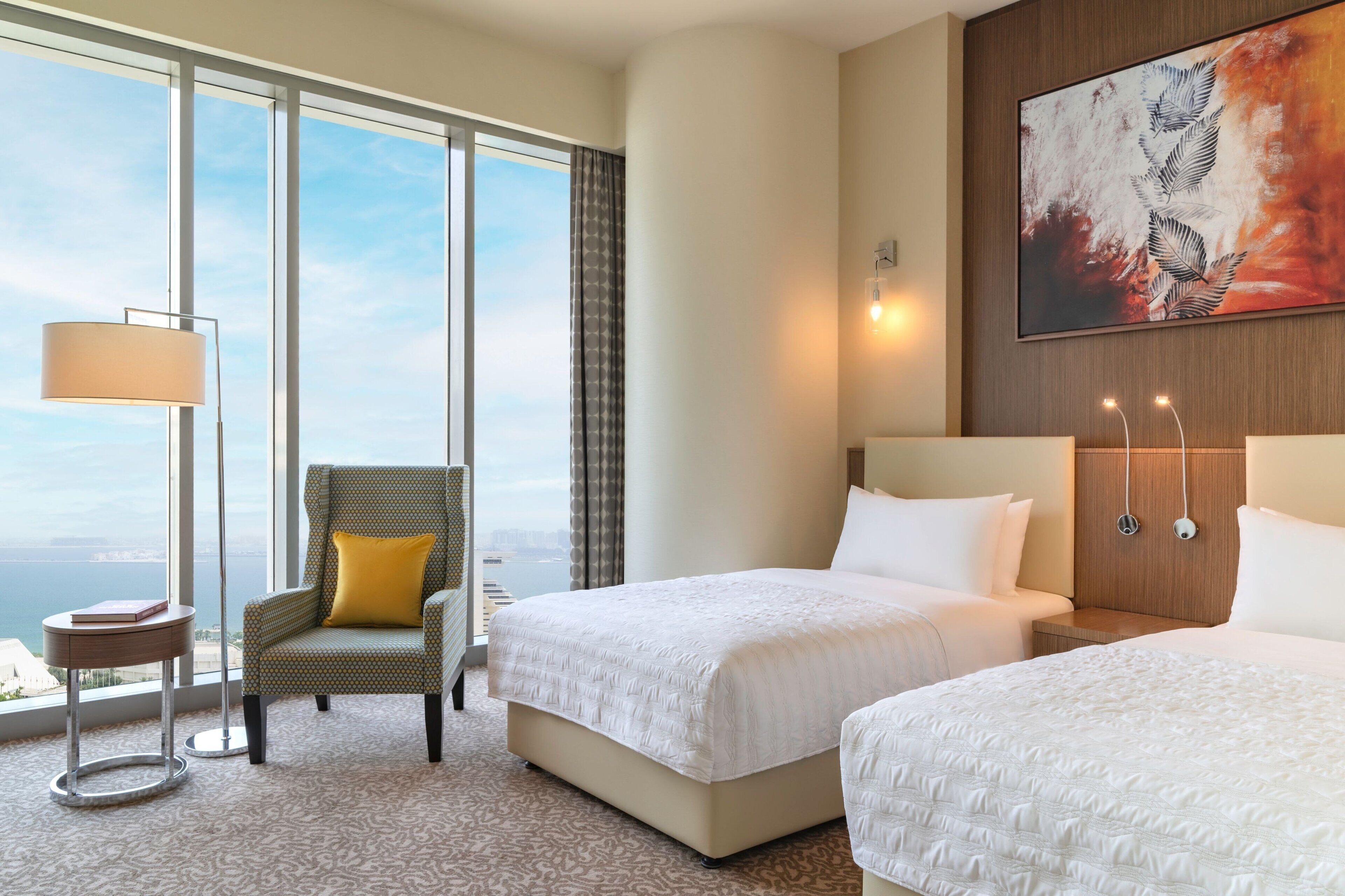 A hotel room with two beds, a chair with a yellow pillow, a side table, a lamp, and a large window offering a view of the city.