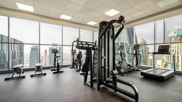 Un gimnasio con diverso equipamiento de ejercicio, ventanas amplias y vistas a un paisaje urbano.